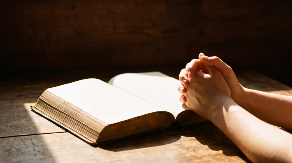 Hands folded in prayer beside an open Bible in warm light